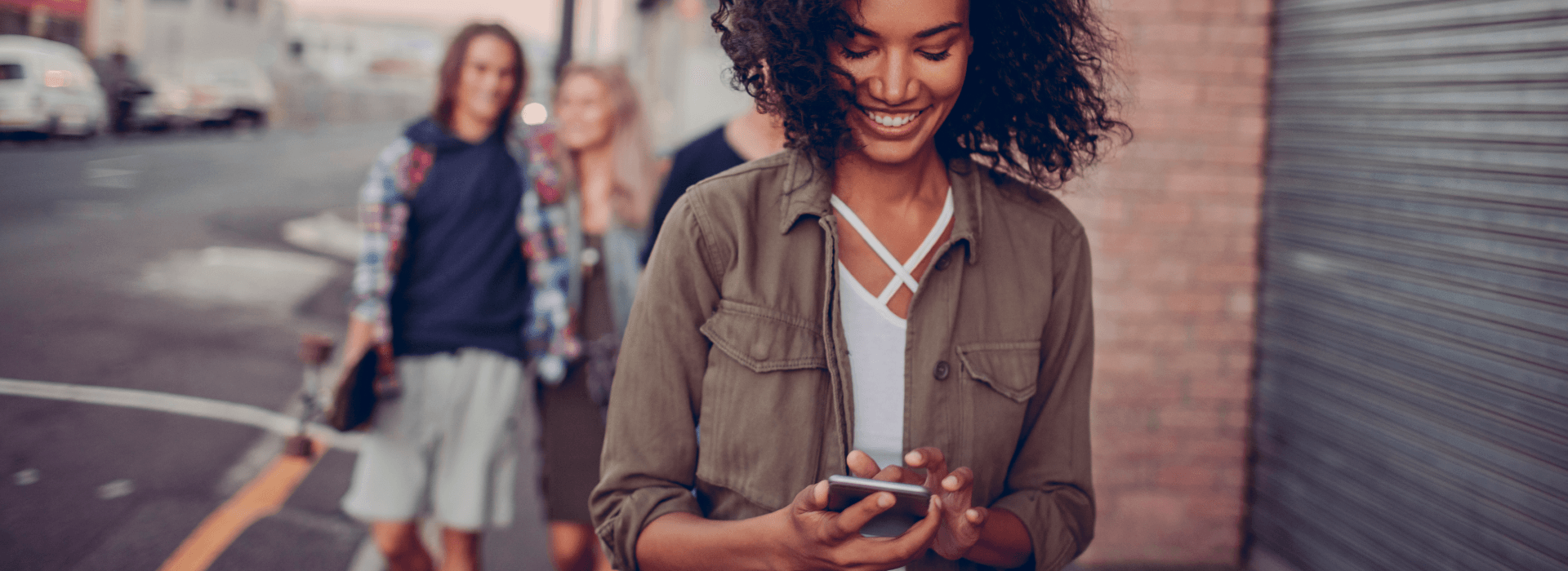 woman looking at her phone , smiling, while walking on the sidewalk with people behind her